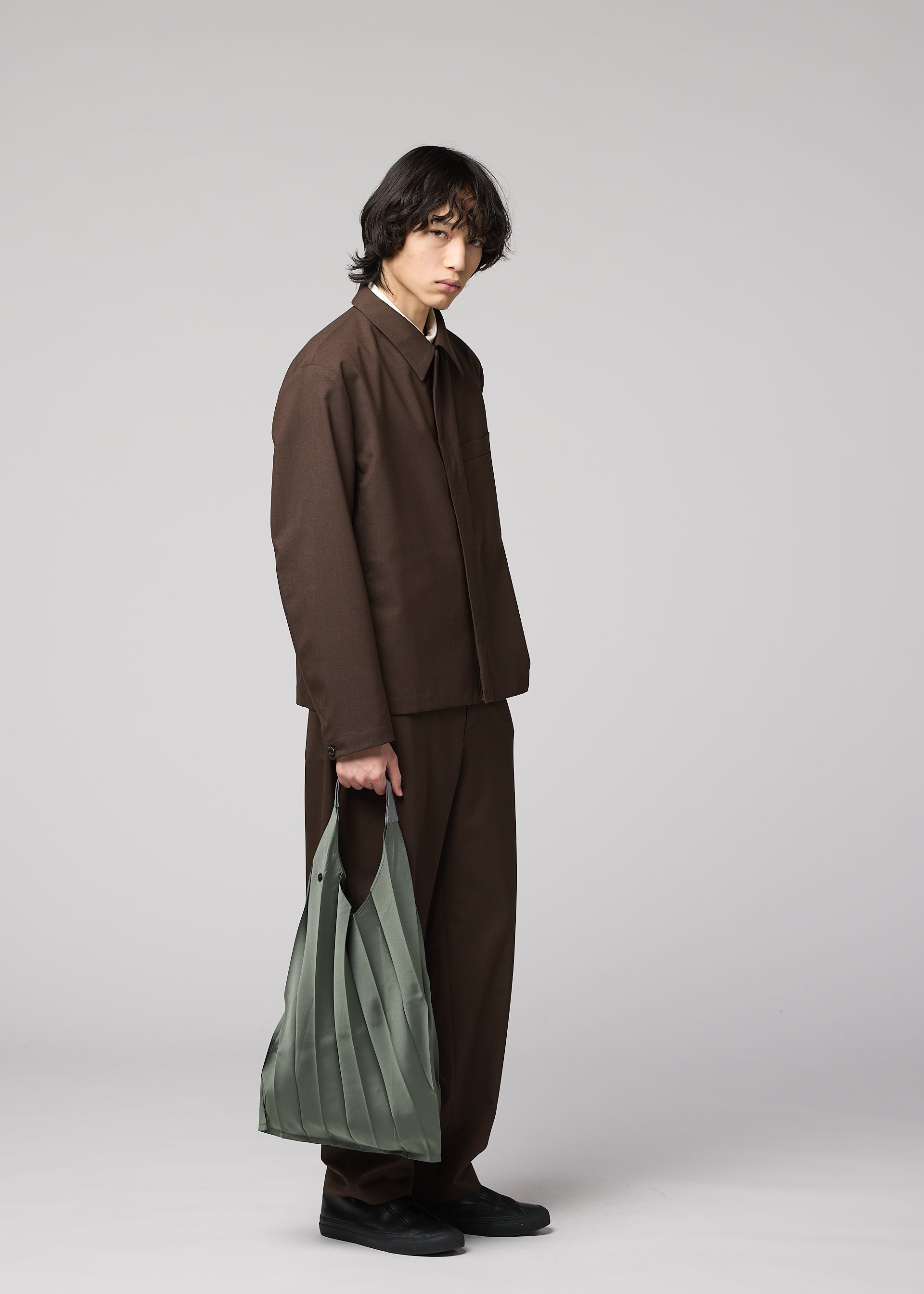 Zoomed view A person with medium-length dark hair stands against a plain light background, wearing a loose brown suit and black shoes, holding the light green GURU GURU foldable tote bag with handmade pleats by IM MEN in their left hand.