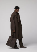 A person in a long dark brown coat, dark pants, and shoes stands against a plain background, holding an IM MEN PATA PA foldable tote bag made from polyester suede by their side.