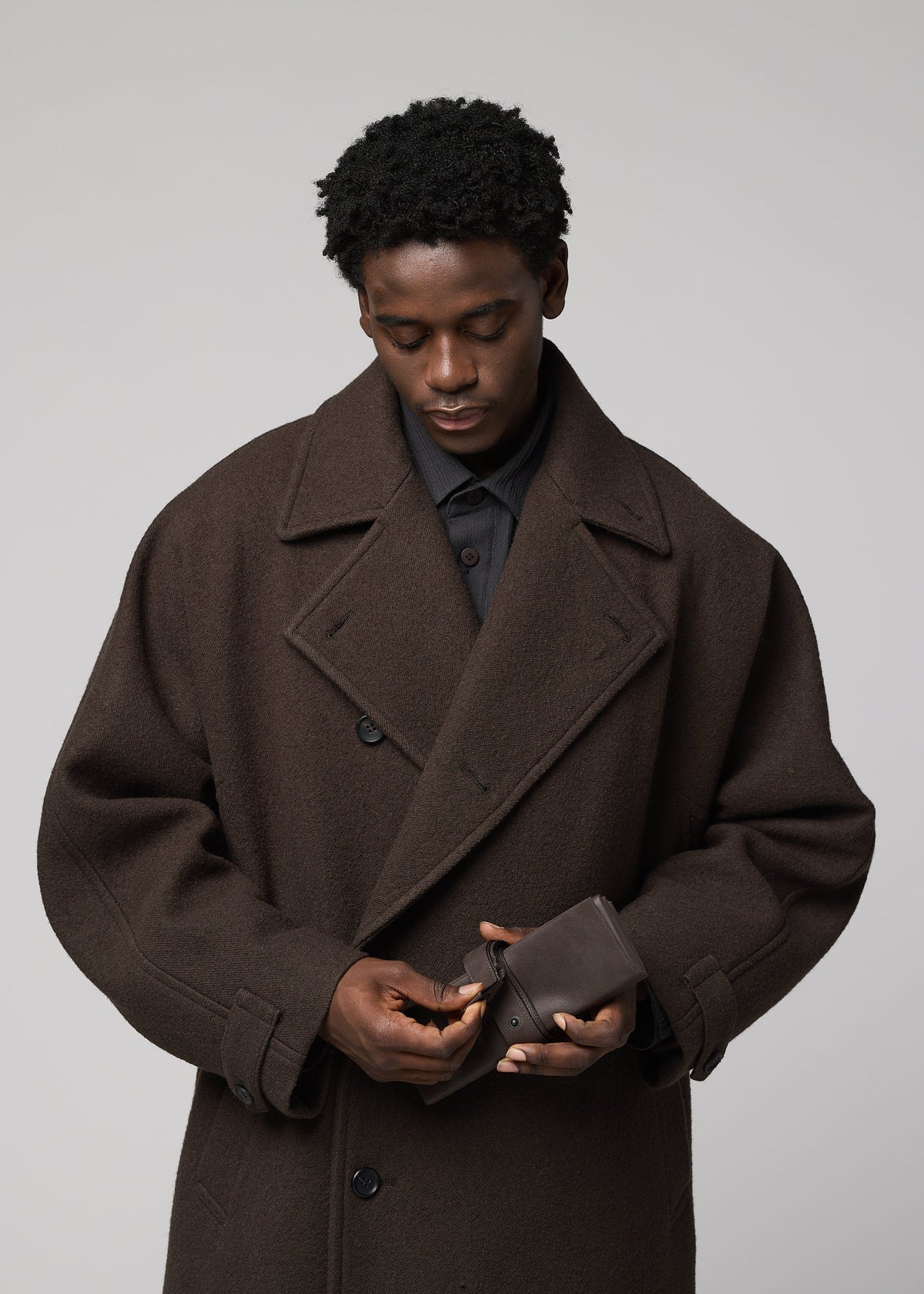 Wearing a dark brown oversized coat, a person looks down as they adjust the IM MEN PATA PA small polyester suede bag against a plain light background. view 9 of 12