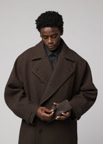 Wearing a dark brown oversized coat, a person looks down as they adjust the IM MEN PATA PA small polyester suede bag against a plain light background.