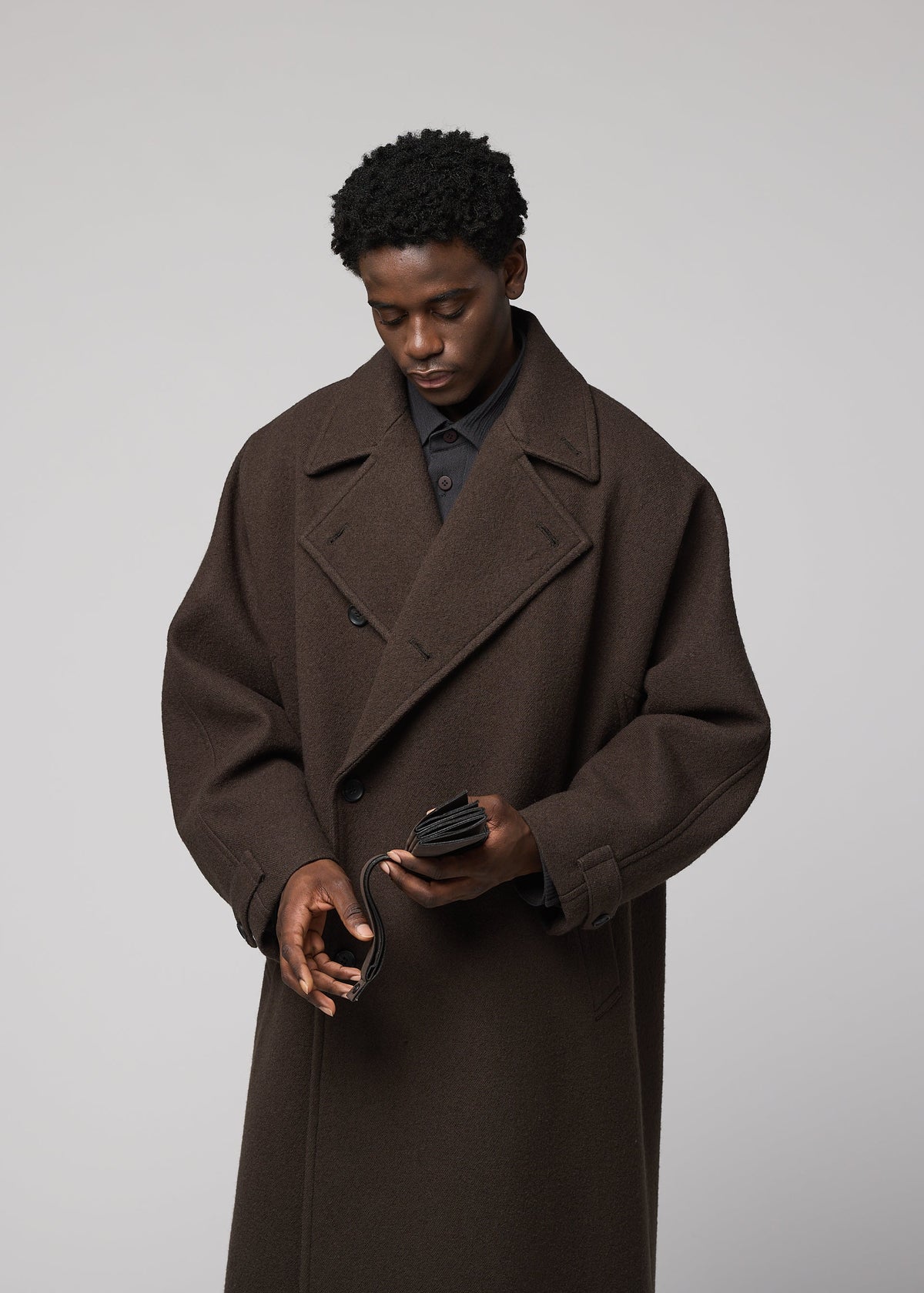 A person in a dark brown oversized coat opens a black PATA PA wallet by IM MEN, with a foldable tote bag nearby, against a plain light gray background. view 10 of 12