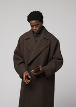 A person in a dark brown oversized coat opens a black PATA PA wallet by IM MEN, with a foldable tote bag nearby, against a plain light gray background.