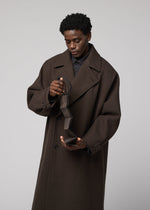A person in a long brown coat looks down, opening an empty wallet beside the PATA PA foldable tote bag by IM MEN, against a plain grey background.