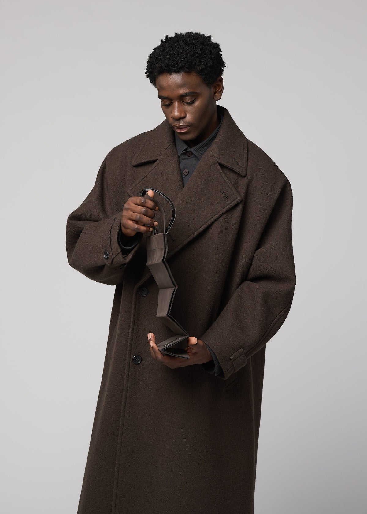 Zoomed view A person in a long brown coat looks down, opening an empty wallet beside the PATA PA foldable tote bag by IM MEN, against a plain grey background.