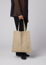 A person in black pants and a dark shirt holds an IM MEN PATA PA foldable tote bag with horizontal stripes and two handles, standing against a plain light background.
