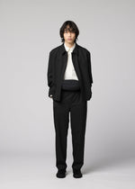 A person with medium-length dark hair stands against a plain background, wearing a black jacket and pants, a white shirt, black shoes, and the IM MEN CUMMERBUND BAG in compact size around the waist. Their hands are in their pockets and their expression is neutral.