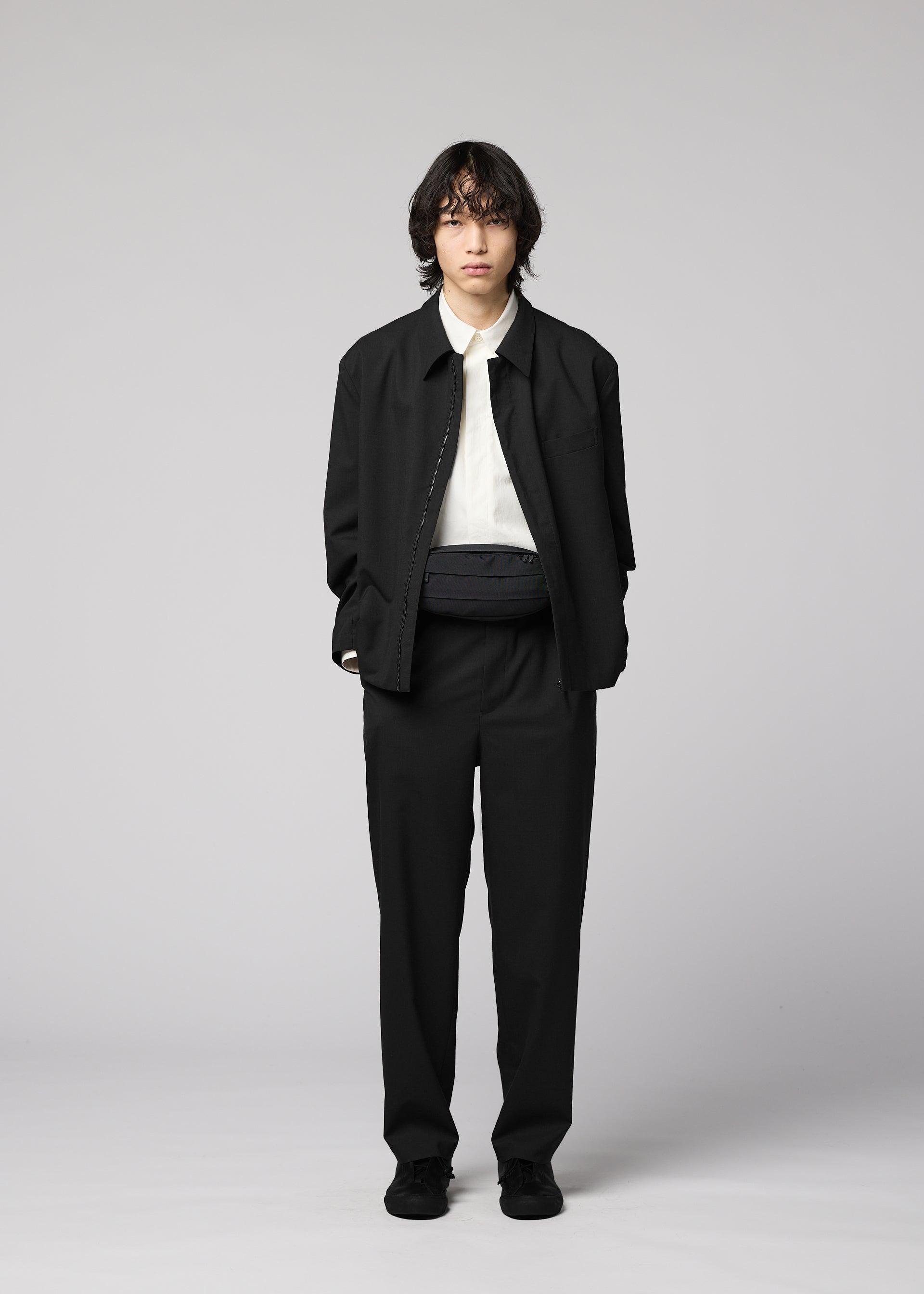 Zoomed view A person with medium-length dark hair stands against a plain background, wearing a black jacket and pants, a white shirt, black shoes, and the IM MEN CUMMERBUND BAG in compact size around the waist. Their hands are in their pockets and their expression is neutral.