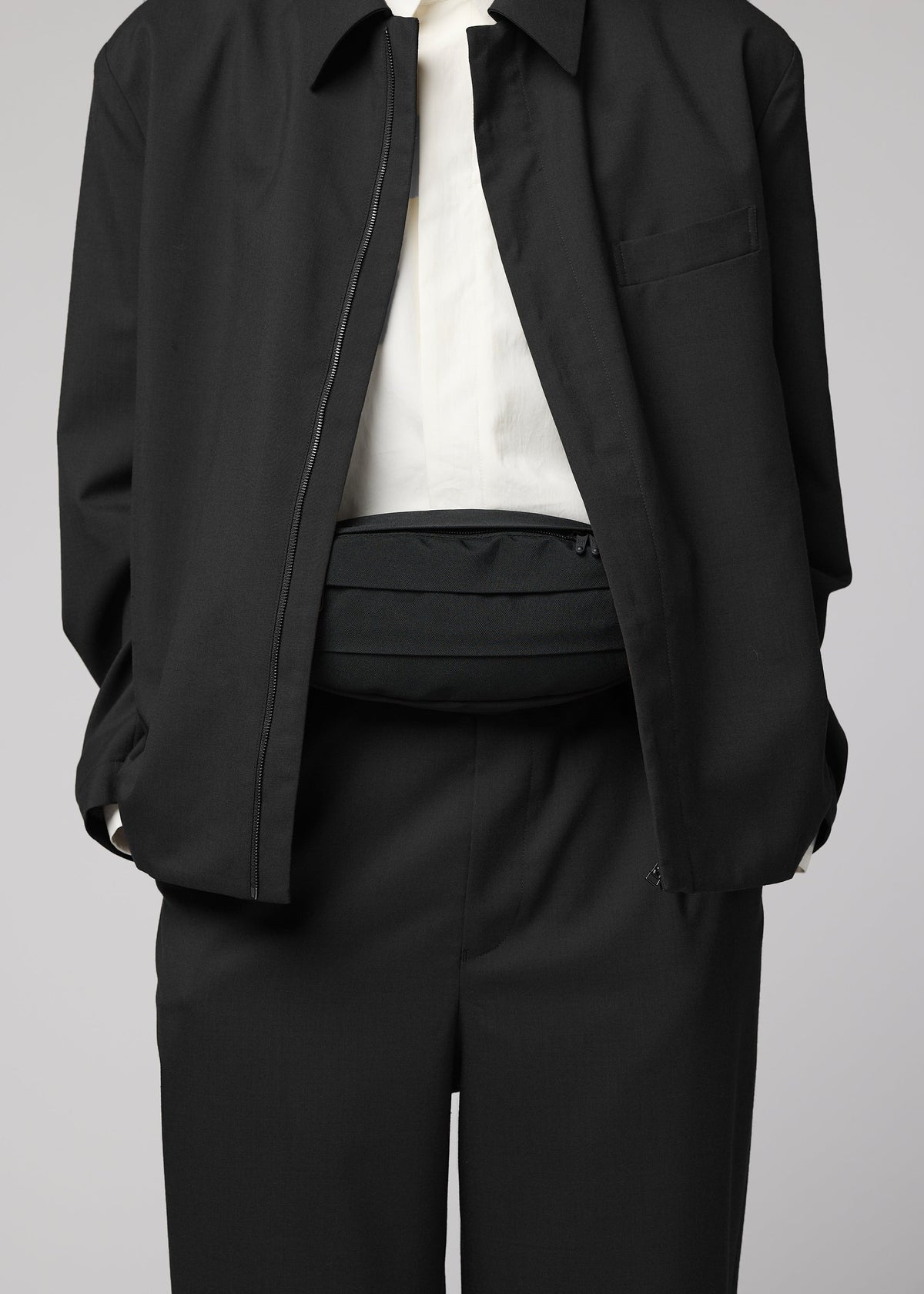 A person dressed in a black suit with a white shirt, jacket unzipped to reveal the IM MEN CUMMERBUND BAG—a compact black belt bag—worn around their waist. The person’s head is not visible. view 9 of 11
