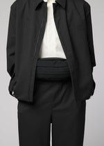 A person dressed in a black suit with a white shirt, jacket unzipped to reveal the IM MEN CUMMERBUND BAG—a compact black belt bag—worn around their waist. The person’s head is not visible.