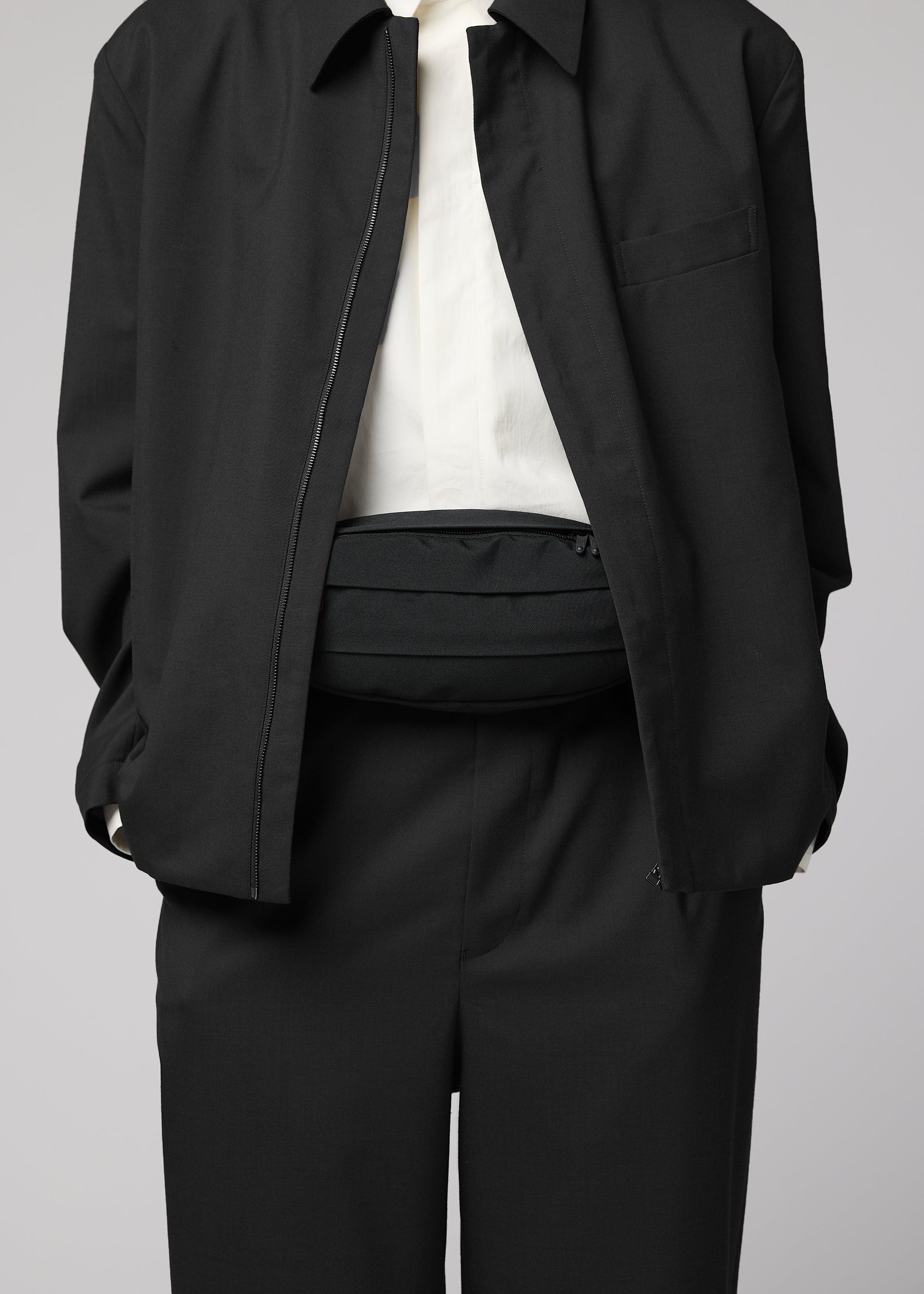 Zoomed view A person dressed in a black suit with a white shirt, jacket unzipped to reveal the IM MEN CUMMERBUND BAG—a compact black belt bag—worn around their waist. The person’s head is not visible.