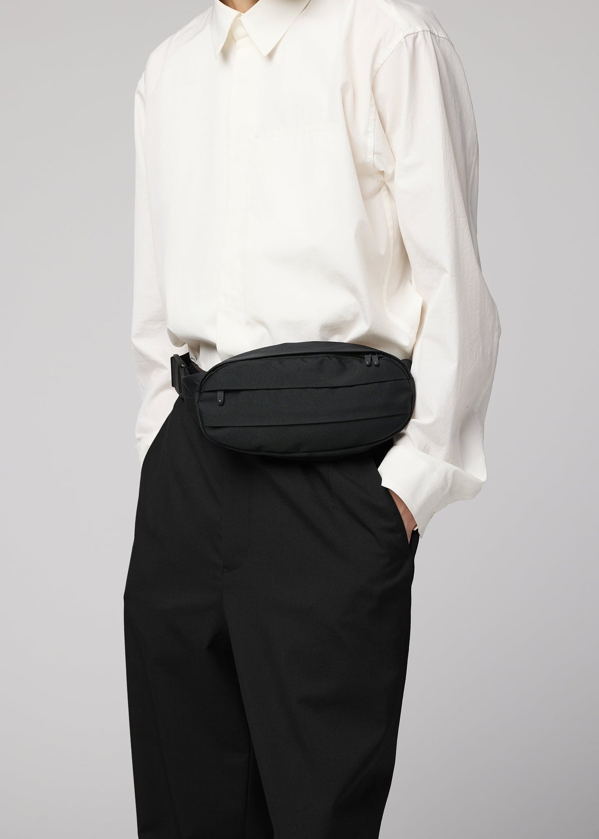 A person wearing a white button-up shirt, black pants, and the IM MEN CUMMERBUND BAG—a compact black waist bag featuring a subtle cummerbund motif—stands with one hand in their pocket. Their head is not visible in the image. view 10 of 11