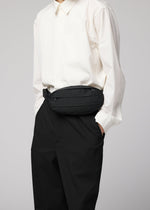 A person wearing a white button-up shirt, black pants, and the IM MEN CUMMERBUND BAG—a compact black waist bag featuring a subtle cummerbund motif—stands with one hand in their pocket. Their head is not visible in the image.