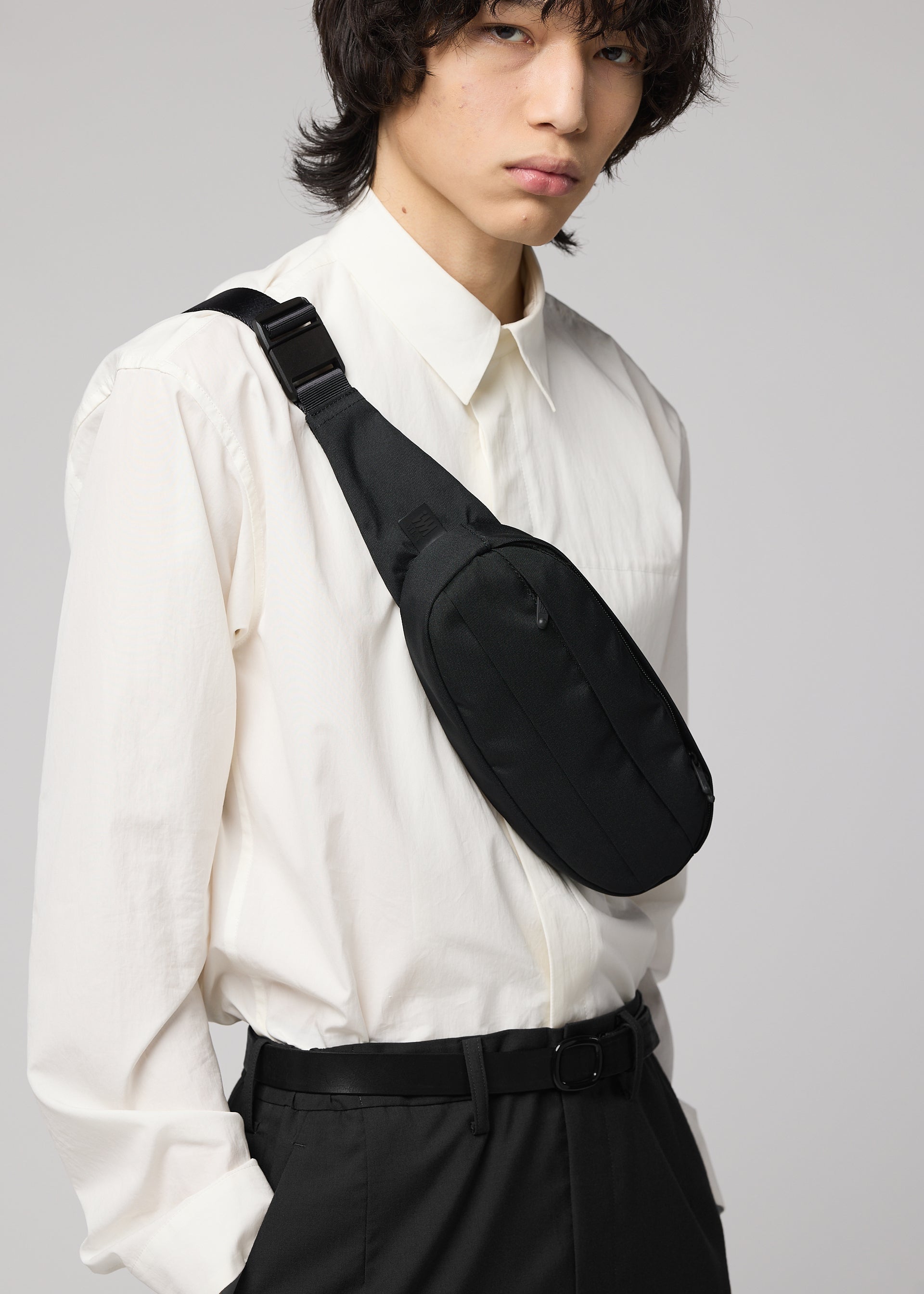 Zoomed view A person stands against a plain light gray background wearing a white button-up shirt, black belt, pants, and the IM MEN CUMMERBUND BAG—a compact black crossbody bag—strapped across their chest.