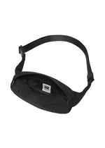Introducing the CUMMERBUND BAG by IM MEN: This sleek black waist bag features an open zipper showcasing its spacious inner compartment and a white tag with black text. The adjustable strap ensures a comfortable fit, while its compact design is ideal for carrying your essentials. Subtle stitching inspired by a cummerbund motif adds an elegant touch to this sophisticated and practical accessory.