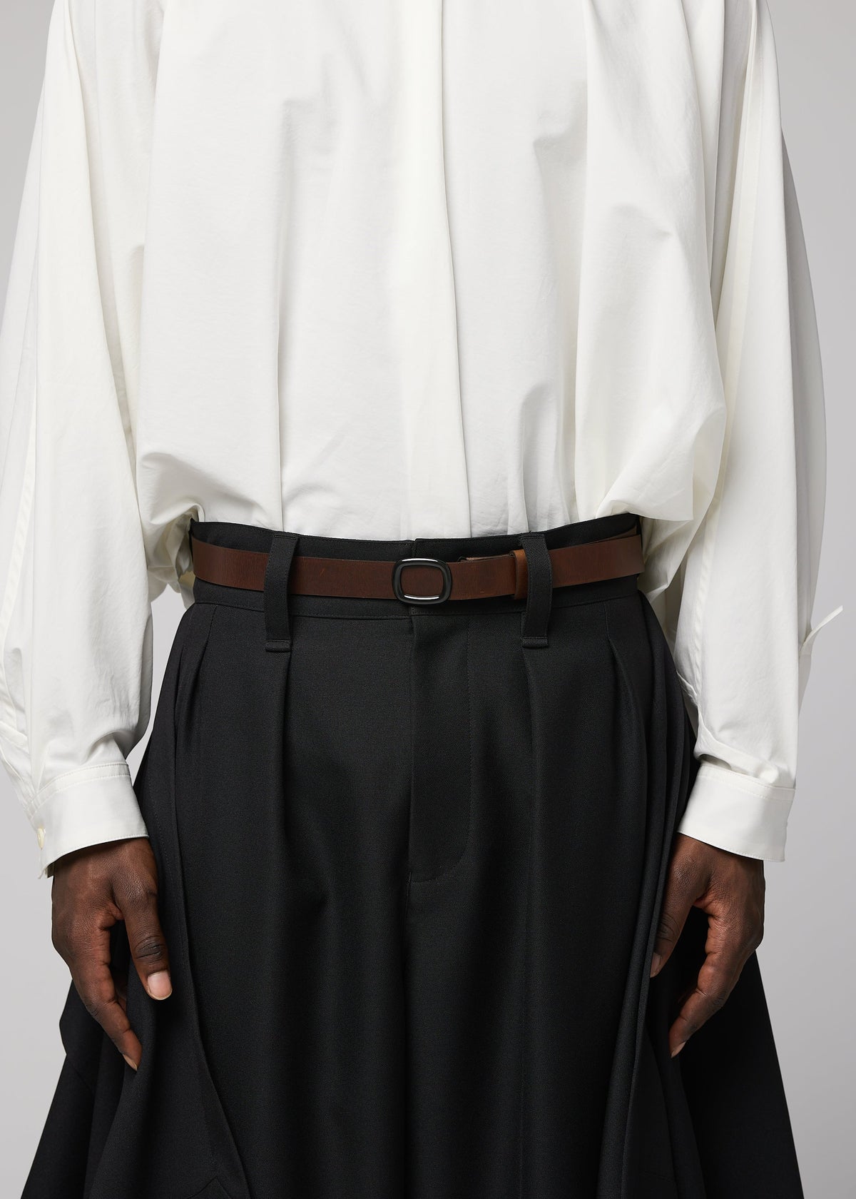 A person is wearing IM MEN’s OVAL loose white shirt tucked into high-waisted, wide-leg black pleated pants, accessorized with an adjustable leather belt featuring an angled buckle. Their hands rest relaxed at their sides. view 6 of 6