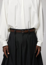 A person is wearing IM MEN’s OVAL loose white shirt tucked into high-waisted, wide-leg black pleated pants, accessorized with an adjustable leather belt featuring an angled buckle. Their hands rest relaxed at their sides.