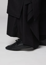 A person wearing the IM MEN FLIPPER long black garment and black lace-up sneakers featuring a cushioned insole stands on a light gray surface.
