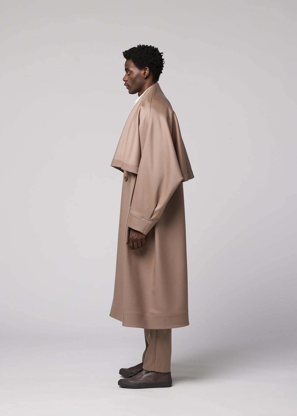 A person stands in profile against a plain gray background, wearing IM MEN’s “FRONT BACK” long, light brown wool coat from the Autumn Winter Collection, paired with matching trousers and dark shoes. view 5 of 9