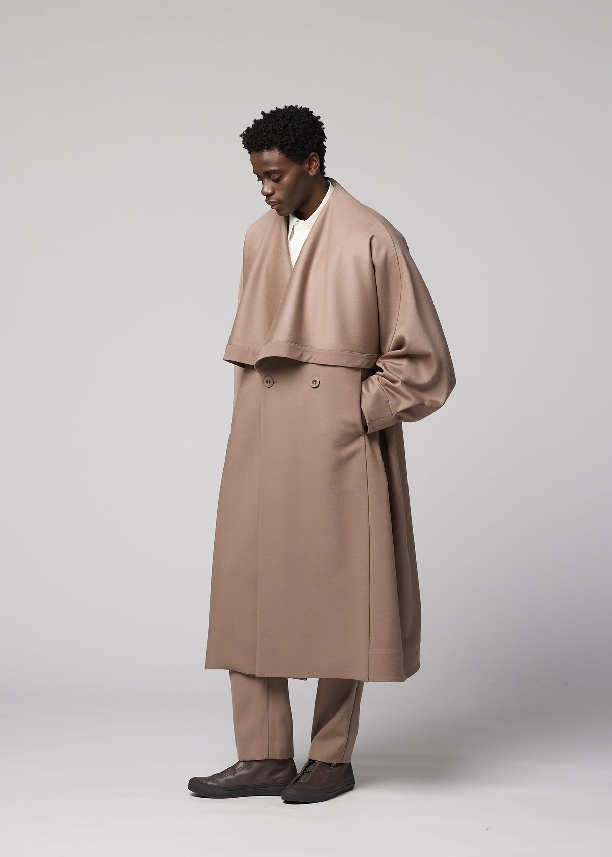 A man stands against a plain backdrop, wearing the IM MEN FRONT BACK long, light brown wool coat with wide lapels and matching trousers from the Autumn Winter Collection. With his hands in his pockets as he looks down, he effortlessly showcases the coat’s refined satin back detailing. view 7 of 9