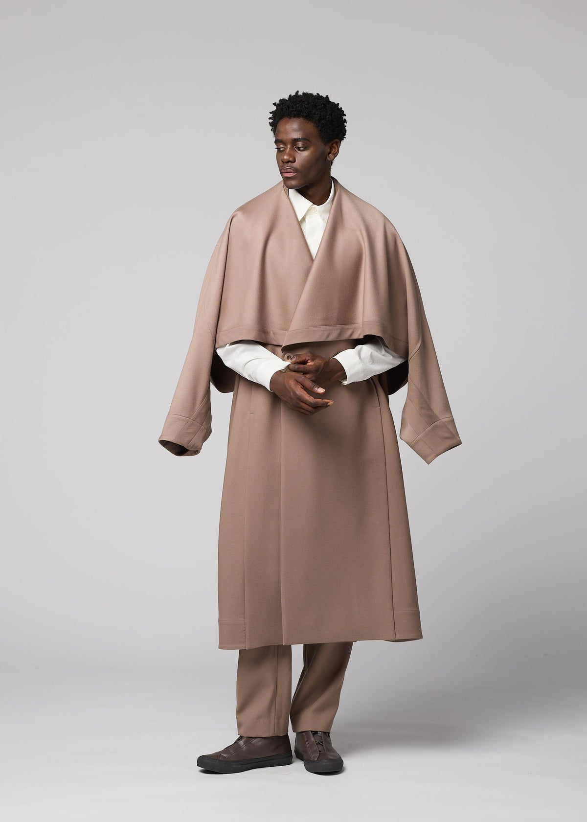 A man stands against a gray backdrop, wearing the oversized beige wool “FRONT BACK” coat with wide sleeves from IM MEN, part of the Autumn Winter Collection. He pairs it with matching beige pants, a white shirt, and dark shoes, looking to the side with his hands loosely clasped. view 8 of 9