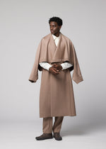 A man stands against a gray backdrop, wearing the oversized beige wool “FRONT BACK” coat with wide sleeves from IM MEN, part of the Autumn Winter Collection. He pairs it with matching beige pants, a white shirt, and dark shoes, looking to the side with his hands loosely clasped.