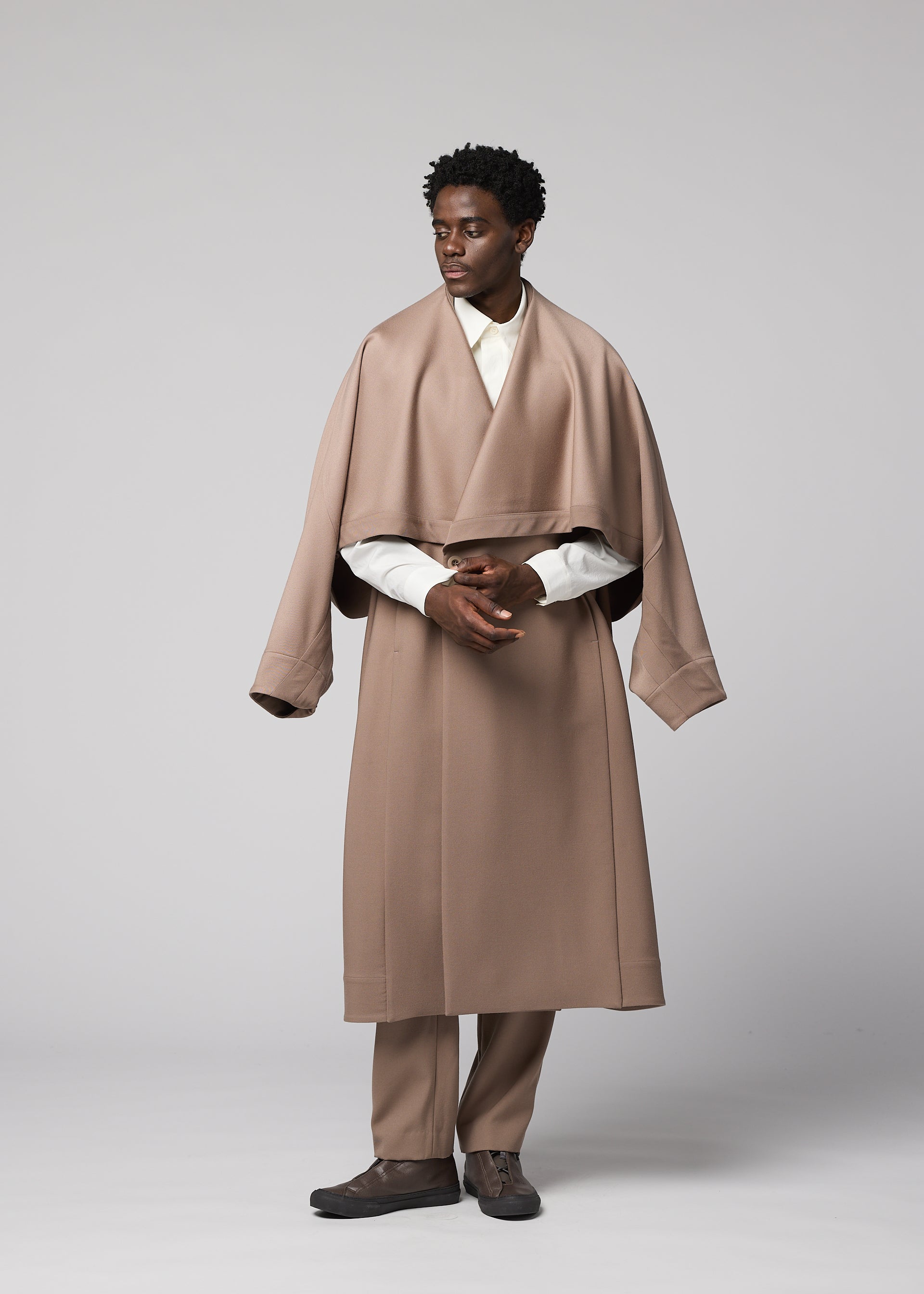 Zoomed view A man stands against a gray backdrop, wearing the oversized beige wool “FRONT BACK” coat with wide sleeves from IM MEN, part of the Autumn Winter Collection. He pairs it with matching beige pants, a white shirt, and dark shoes, looking to the side with his hands loosely clasped.