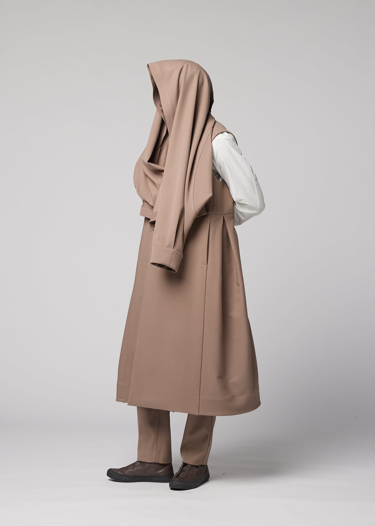 A person stands against a light background wearing the FRONT BACK beige wool coat by IM MEN, with fabric draped over their head to conceal their face and upper body. Their hands are behind their back. view 9 of 9