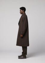 A person with short curly hair stands in profile, wearing the IM MEN FULLER WOOL long dark brown coat, brown pants, and dark shoes against a plain light gray background.