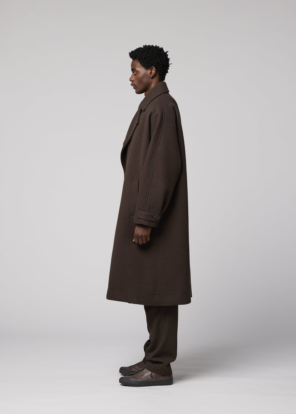 Zoomed view A person with short curly hair stands in profile, wearing the IM MEN FULLER WOOL long dark brown coat, brown pants, and dark shoes against a plain light gray background.