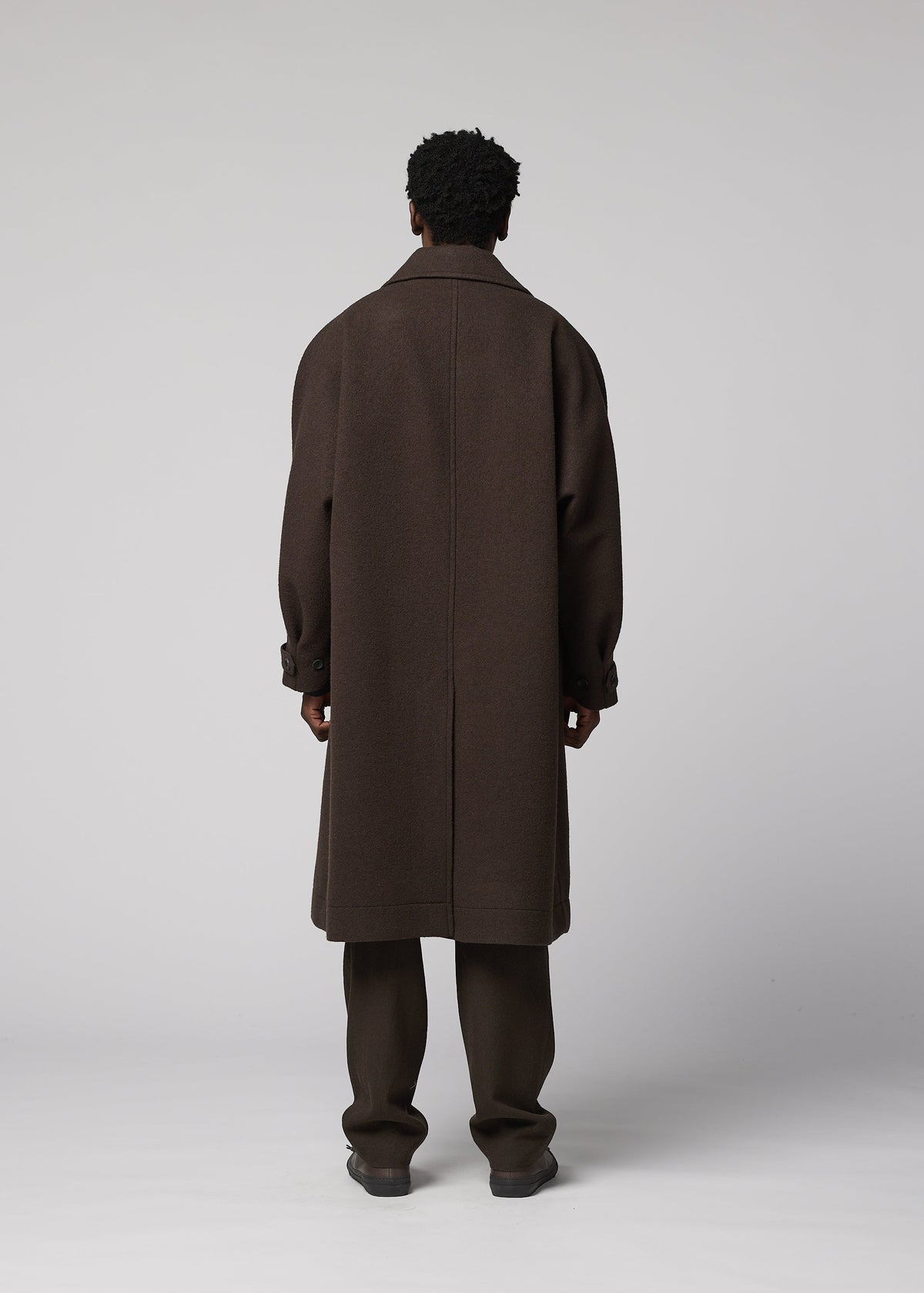 A person with short curly hair faces away, wearing the IM MEN FULLER WOOL long dark brown coat with a standing collar, paired with brown pants and brown shoes against a plain light gray background. view 7 of 8
