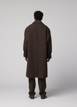 A person with short curly hair faces away, wearing the IM MEN FULLER WOOL long dark brown coat with a standing collar, paired with brown pants and brown shoes against a plain light gray background.
