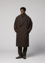 A man stands against a plain backdrop wearing the IM MEN FULLER WOOL coat, paired with brown pants and dark shoes. His hands are in his pockets as he looks slightly to the side.