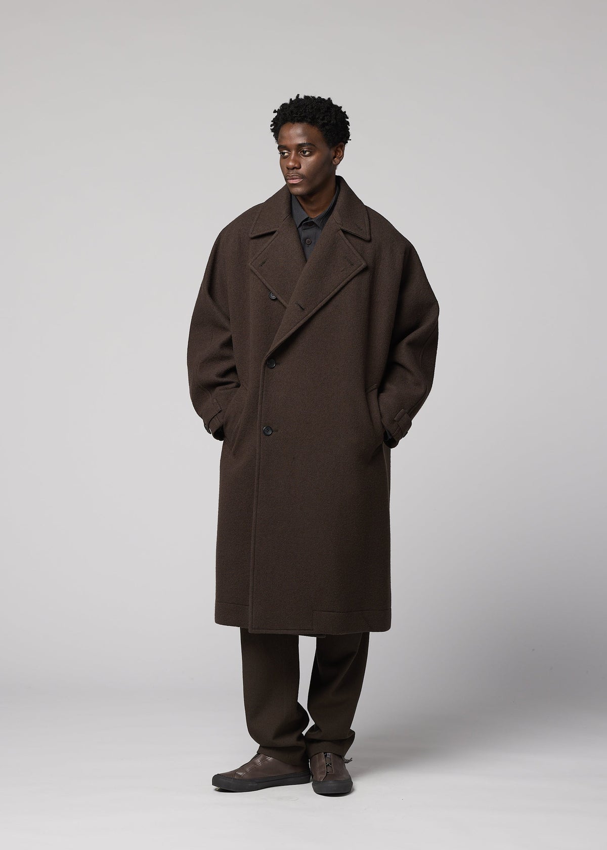 Zoomed view A man stands against a plain backdrop wearing the IM MEN FULLER WOOL coat, paired with brown pants and dark shoes. His hands are in his pockets as he looks slightly to the side.