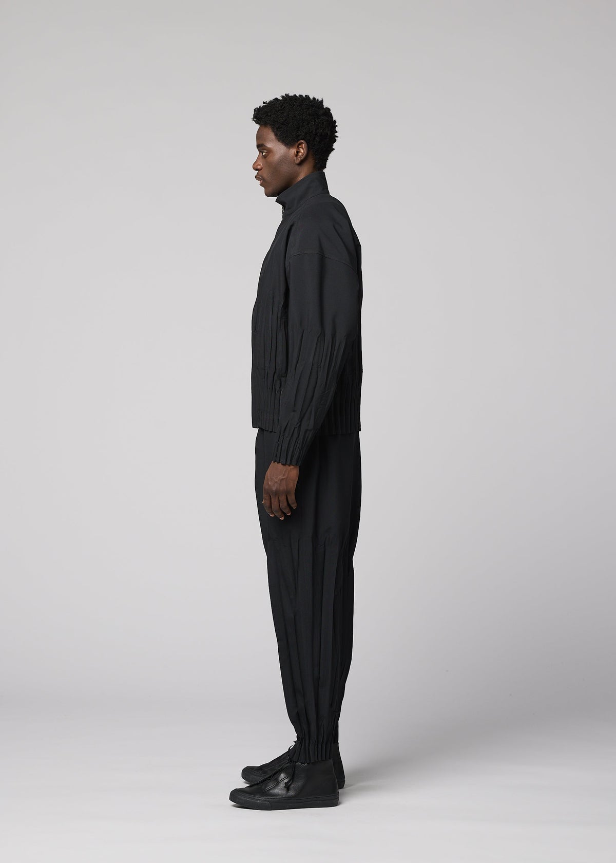 A person stands in profile facing left, dressed in IM MEN’s WAFT BLACK polyester jacket with a subtle linen-like texture, black pants, and black shoes against a plain light gray background. view 5 of 8