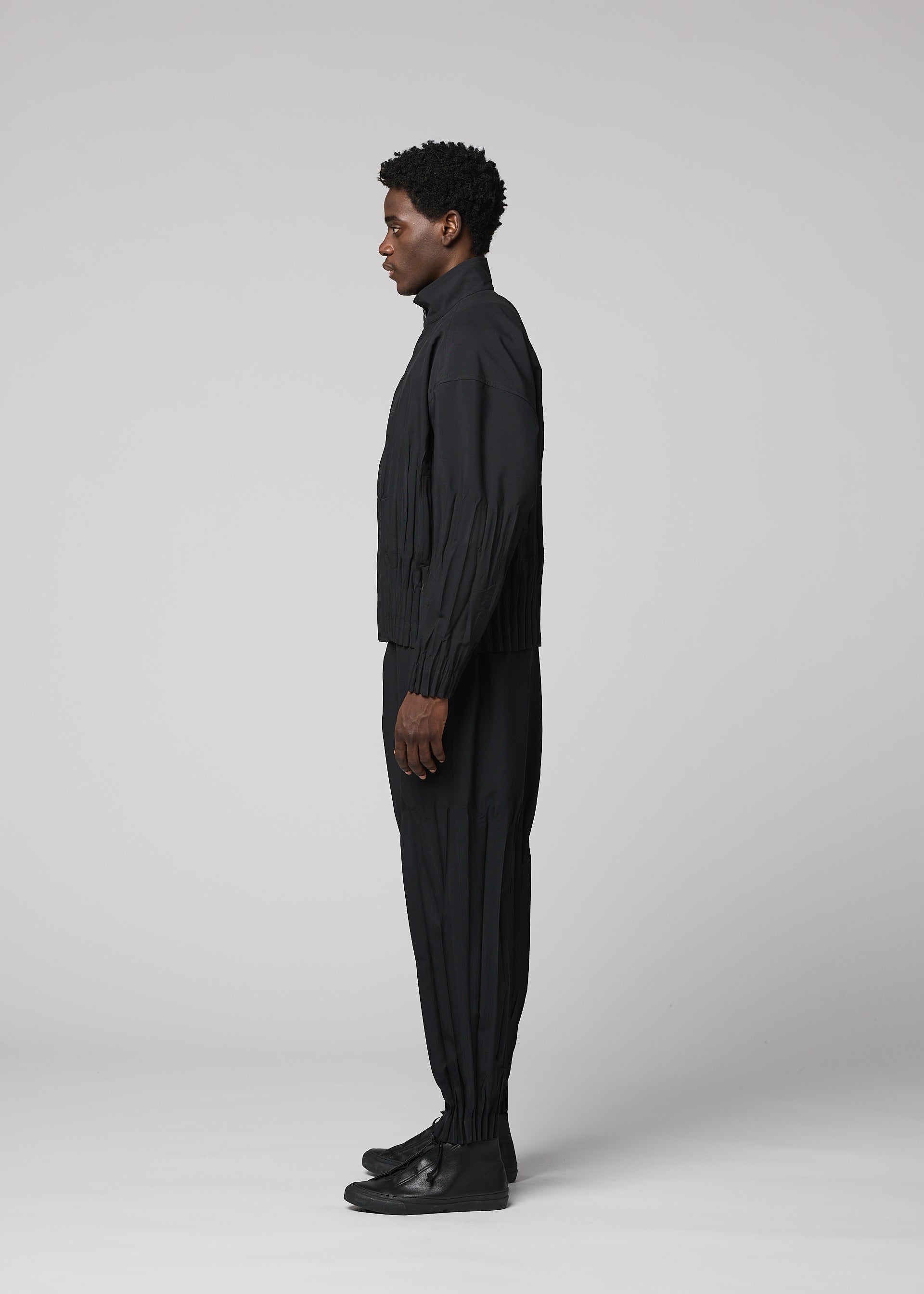 Zoomed view A person stands in profile facing left, dressed in IM MEN’s WAFT BLACK polyester jacket with a subtle linen-like texture, black pants, and black shoes against a plain light gray background.