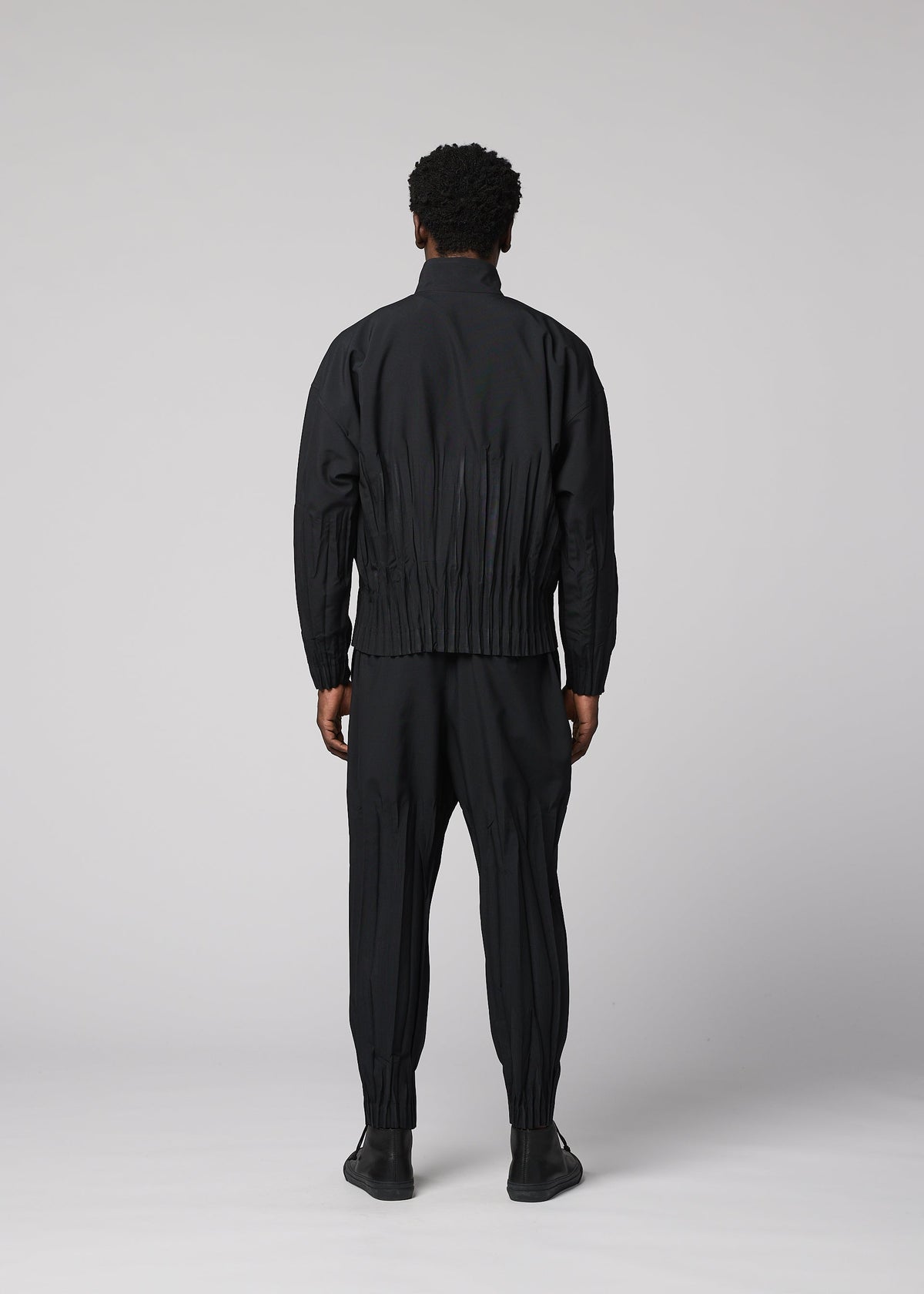 A person faces away from the camera, wearing the WAFT BLACK jacket and pants by IM MEN—both with gathered details and linen-like texture—paired with black shoes against a plain light gray background. view 6 of 8
