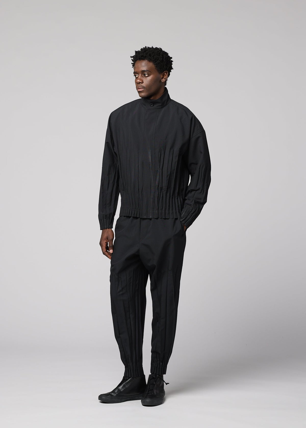 A person stands against a light gray background, wearing the IM MEN WAFT BLACK set: a loose-fitting polyester jacket and matching pleated black pants with a linen-like texture, paired with black shoes. They look slightly to the side with a neutral expression. view 7 of 8