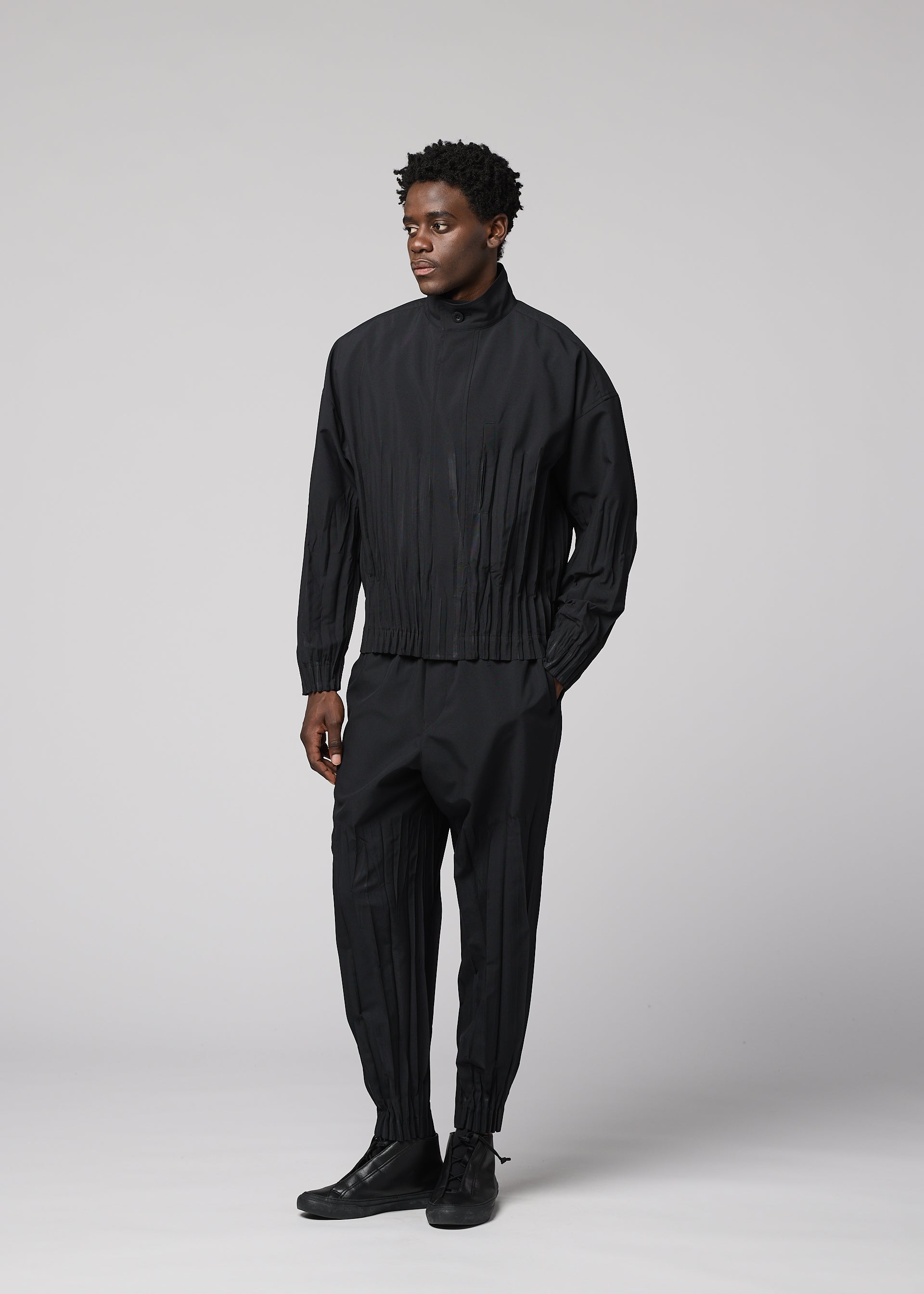 Zoomed view A person stands against a light gray background, wearing the IM MEN WAFT BLACK set: a loose-fitting polyester jacket and matching pleated black pants with a linen-like texture, paired with black shoes. They look slightly to the side with a neutral expression.