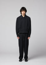 A person with medium-length dark hair stands facing forward in a plain studio setting with a light gray background, wearing the IM MEN COMFORT COTTON jacket—crafted from high-twist organic cotton for a crisp texture and subtle structure—paired with matching black pants and black shoes, creating a refined minimalist look.