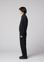 A person with medium-length dark hair stands in profile, facing left, dressed in IM MEN’s COMFORT COTTON black collared jacket made from high-twist cotton for a crisp texture, paired with matching black pants and black shoes against a plain light gray background.