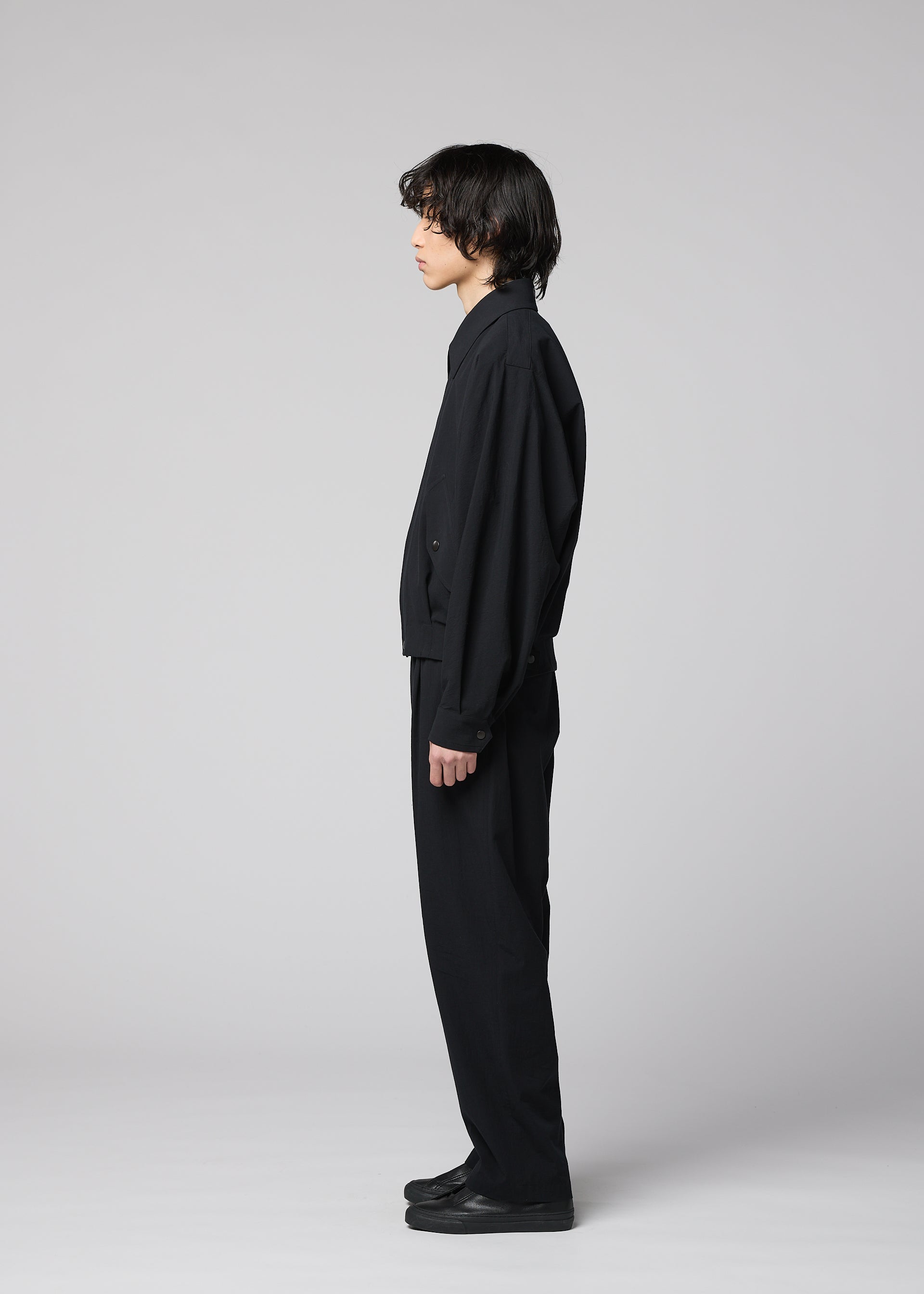 Zoomed view A person with medium-length dark hair stands in profile, facing left, dressed in IM MEN’s COMFORT COTTON black collared jacket made from high-twist cotton for a crisp texture, paired with matching black pants and black shoes against a plain light gray background.