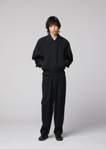 A person stands against a plain light background, dressed in IM MEN’s COMFORT COTTON loose-fitting black jacket made from crisp organic cotton, paired with matching black pants and black shoes. Their hands are in their pockets as they look forward with a neutral expression.