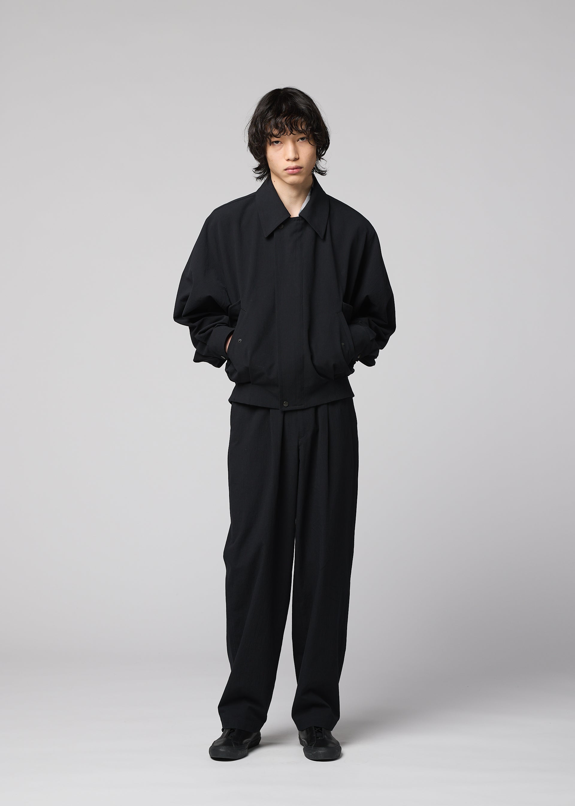 Zoomed view A person stands against a plain light background, dressed in IM MEN’s COMFORT COTTON loose-fitting black jacket made from crisp organic cotton, paired with matching black pants and black shoes. Their hands are in their pockets as they look forward with a neutral expression.