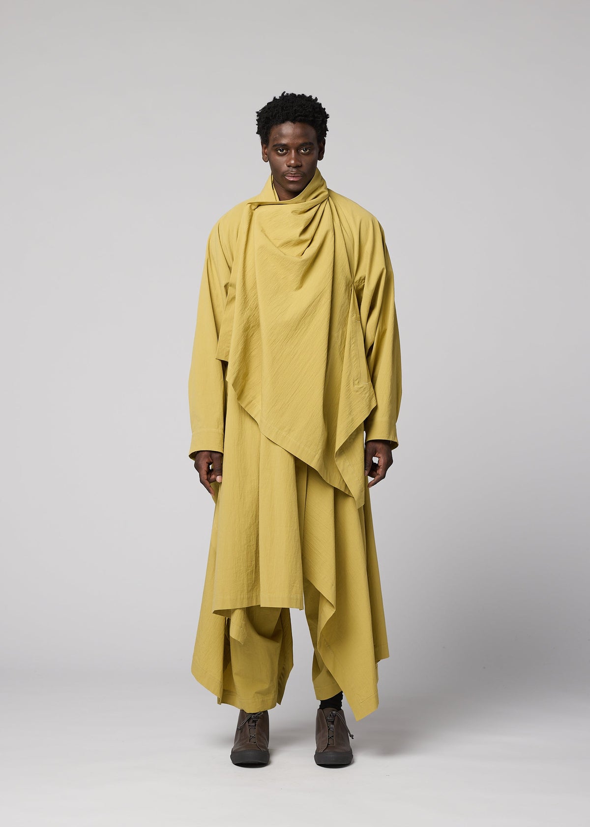 A person stands facing forward against a plain, light gray background, dressed in layered mustard yellow clothing from IM MEN. The outfit features the COMFORT COTTON ensemble, including matching pants and brown shoes. The look is highlighted by a distinctive irregular-cut jacket made from high-twist organic cotton for comfort and versatile styling. view 5 of 9