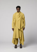A person stands facing forward against a plain, light gray background, dressed in layered mustard yellow clothing from IM MEN. The outfit features the COMFORT COTTON ensemble, including matching pants and brown shoes. The look is highlighted by a distinctive irregular-cut jacket made from high-twist organic cotton for comfort and versatile styling.