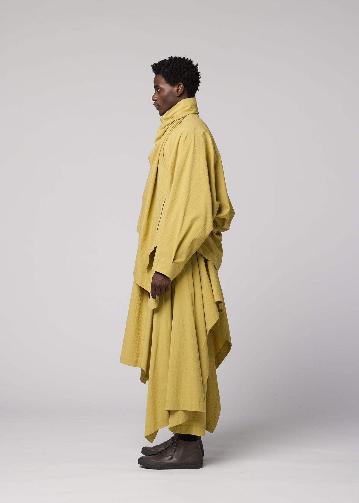 A person standing in profile, dressed in IM MEN’s COMFORT COTTON ensemble—a loose, layered yellow outfit featuring an irregular-cut jacket with a high collar—set against a plain light background. view 6 of 9