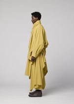 A person standing in profile, dressed in IM MEN’s COMFORT COTTON ensemble—a loose, layered yellow outfit featuring an irregular-cut jacket with a high collar—set against a plain light background.