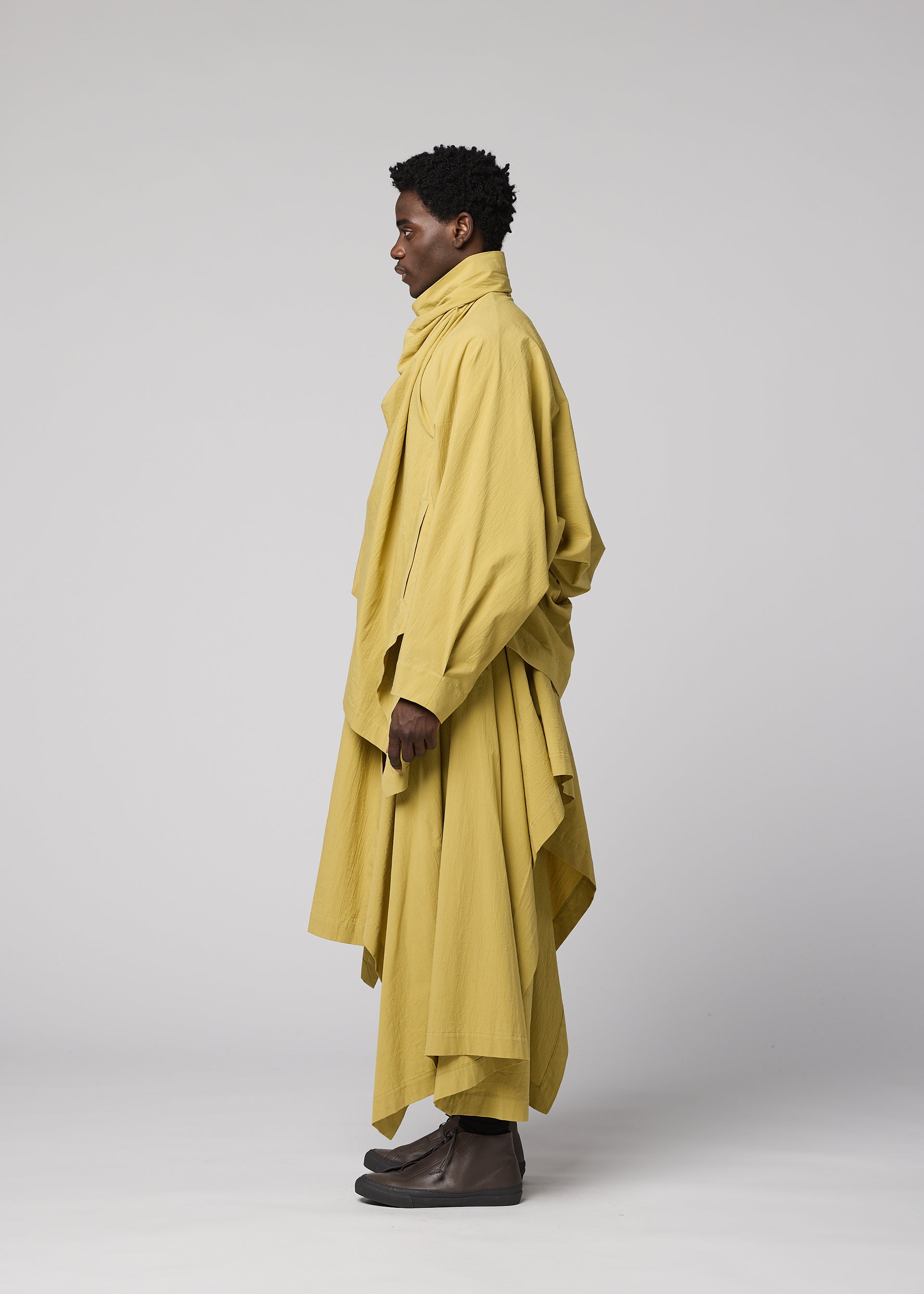 Zoomed view A person standing in profile, dressed in IM MEN’s COMFORT COTTON ensemble—a loose, layered yellow outfit featuring an irregular-cut jacket with a high collar—set against a plain light background.