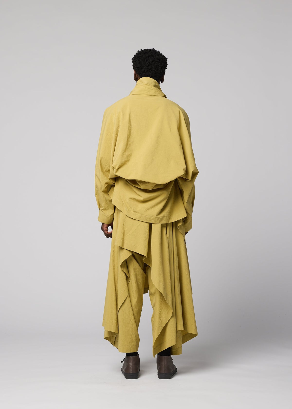 A person stands facing away, dressed in loose, layered yellow clothing with draped fabric and wide pants. The outfit features the COMFORT COTTON irregular-cut jacket by IM MEN, crafted from high-twist organic cotton and demonstrating versatile styling against a plain light gray background. view 7 of 9