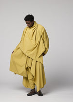 Against a plain backdrop, a person wears loose, layered, oversized yellow clothing from IM MEN’s COMFORT COTTON collection, featuring an irregular-cut jacket made of high-twist organic cotton. With dark shoes and one hand holding the garment, they embody versatile styling while gazing downward thoughtfully.